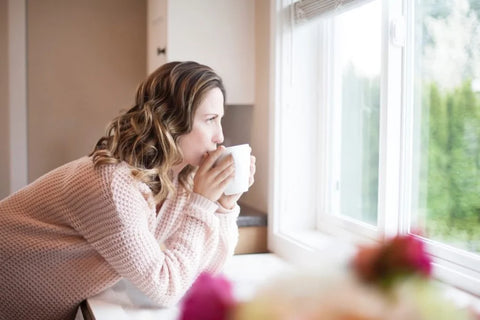 Andrea in a sweater standing by a bright window, drinking from a white mug.