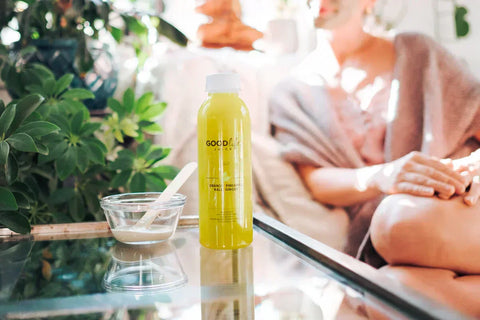 GLJ Glo bottle on a glass table beside a bowl of face mask, surrounded by green plants and Andrea in a robe.