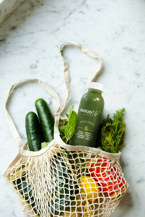 GLJ Detox Greens bottle with accompanying vegetables, fruits, and ingredients in a mesh shopping bag on a marble surface.